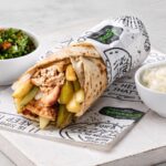 A Lebanese roll meal featuring charcoal chicken, pickles, chips, with side servings of tabouli salad and creamy garlic sauce.